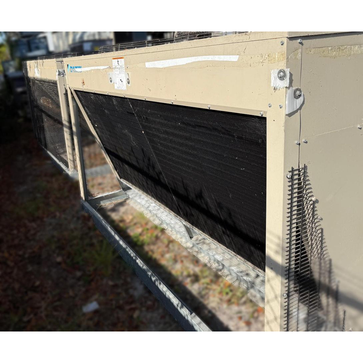 Daikin DPS012AHHE2DW-4 rooftop HVAC unit condenser coil section closeup showing coil fins, wire mesh guard, curb mount base, and beige powder-coated steel cabinet
