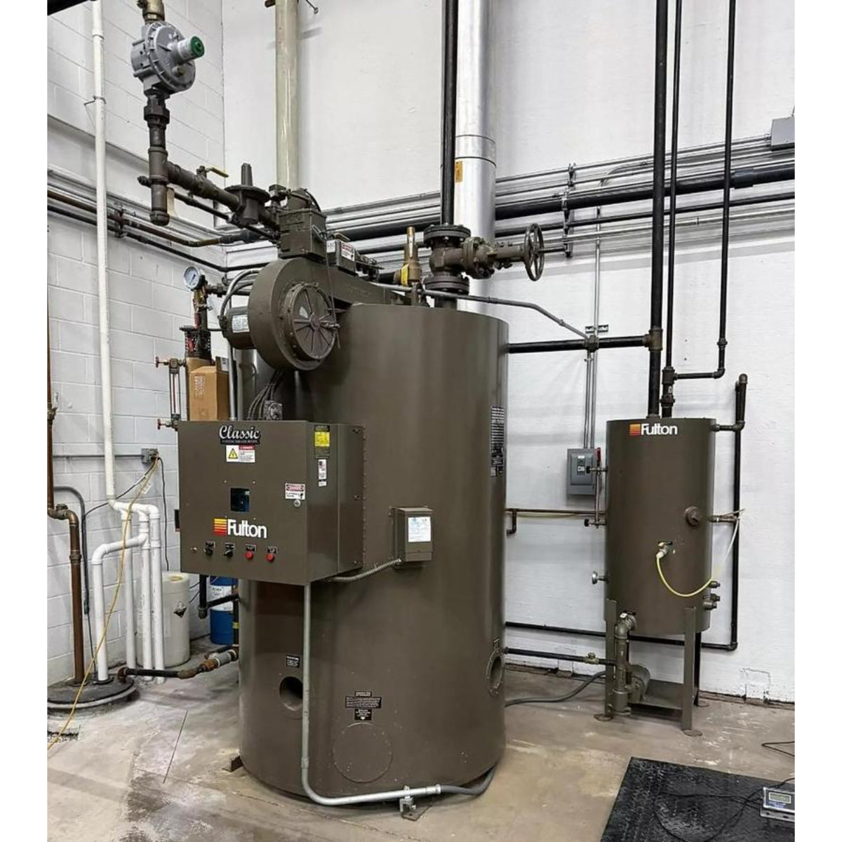 Two Fulton industrial boilers in a mechanical room with pipes and walls.