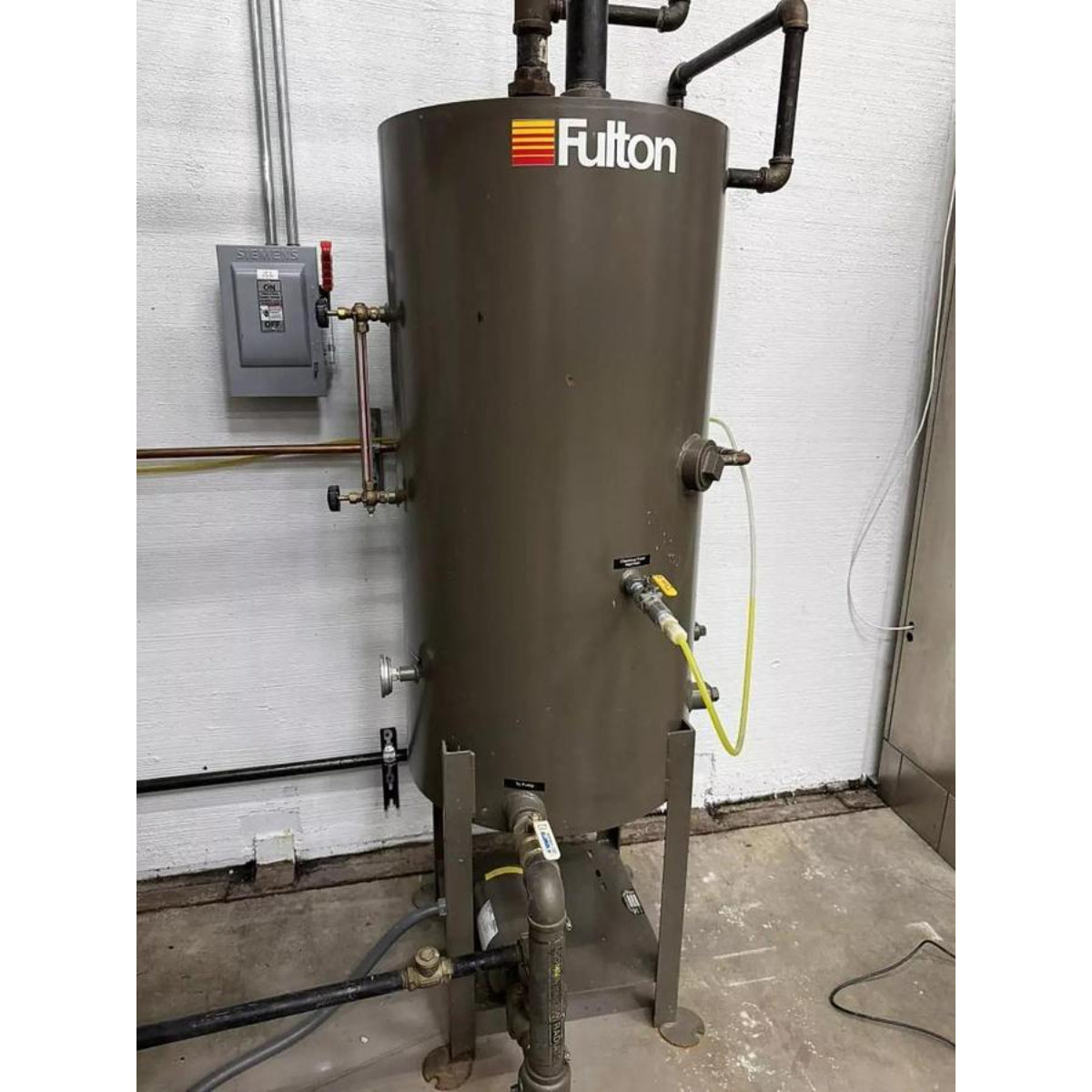 Fulton water treatment tank with pipes and valves against a white wall.