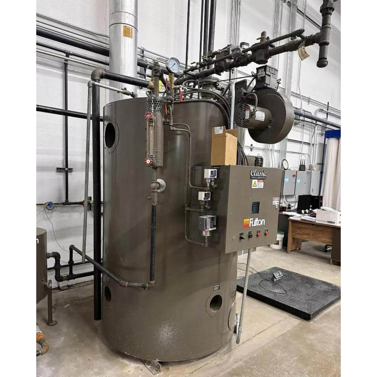Large industrial boiler with various pipes and equipment in a room with large windows.